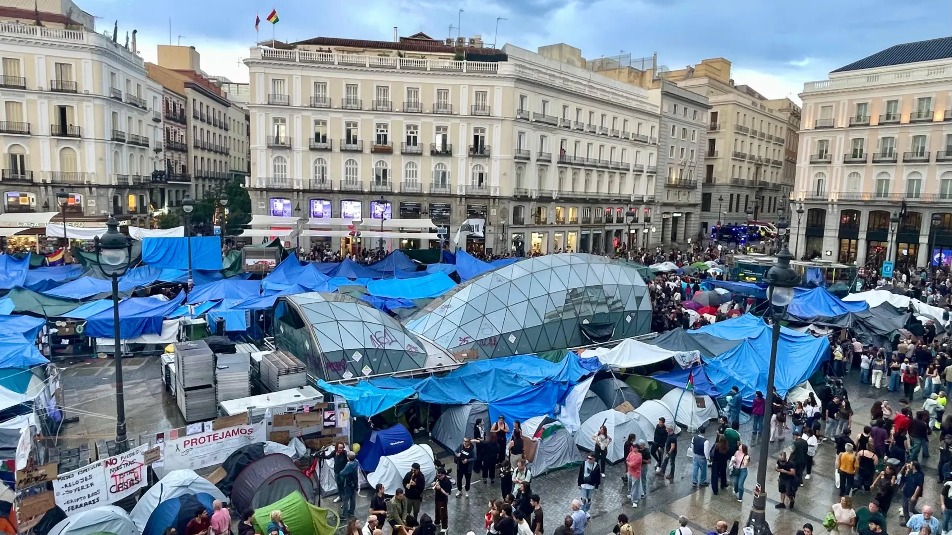 Jóvenes en una calle urbana con carteles de alquiler y propietarios en una reunión.
